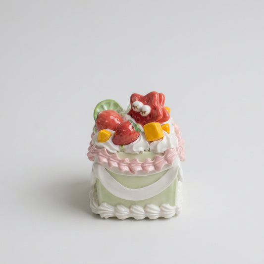 Front view of a pale green ceramic cake with pink piping, white garlands and miniature fruit details.