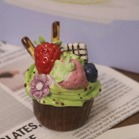 Small dessert with green frosting, strawberries, and blueberries on a wooden stick, placed on a newspaper.